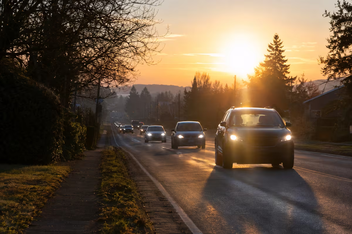 Cars with headlights on drive into a low, glaring sun on a two-lane road at dawn.