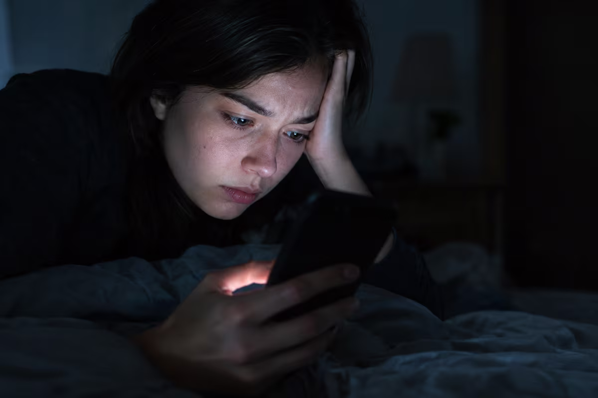 A young woman in a dark bedroom looks at her phone, its glow lighting her worried face.